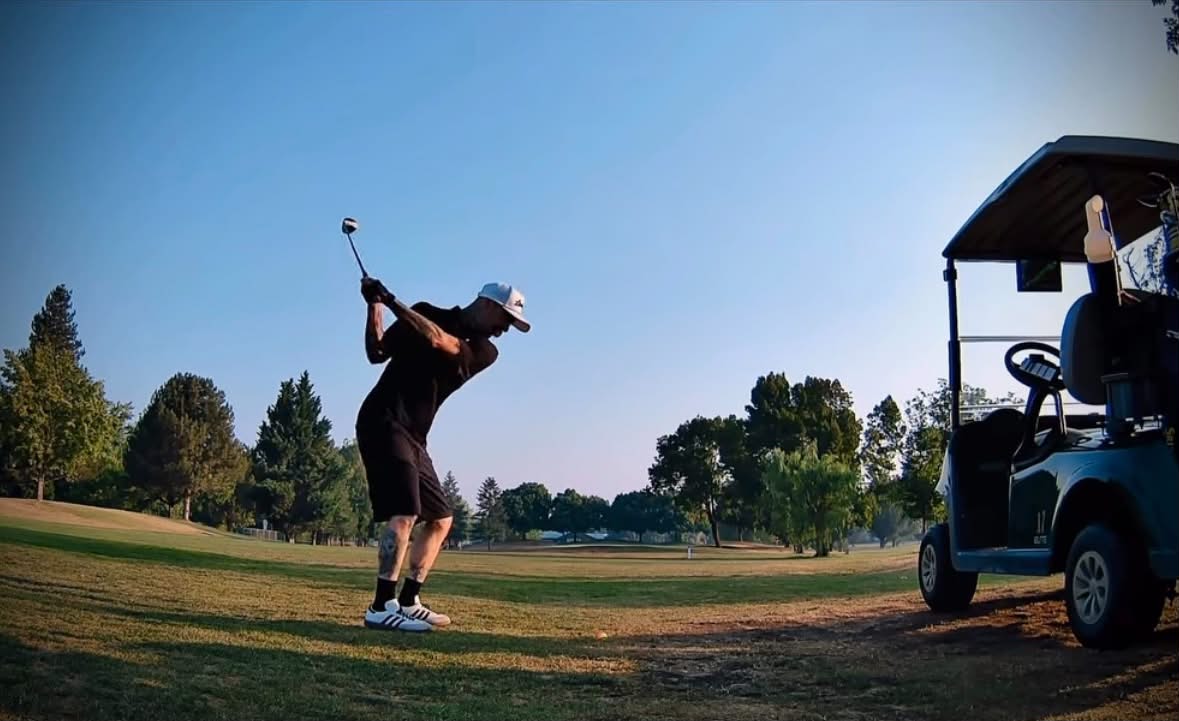 Person swinging a golf club on a golf course with trees and a clear sky in the background.