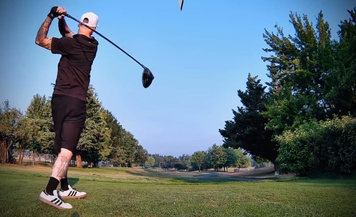 Person swinging a golf club on a golf course with trees and blue sky in background.