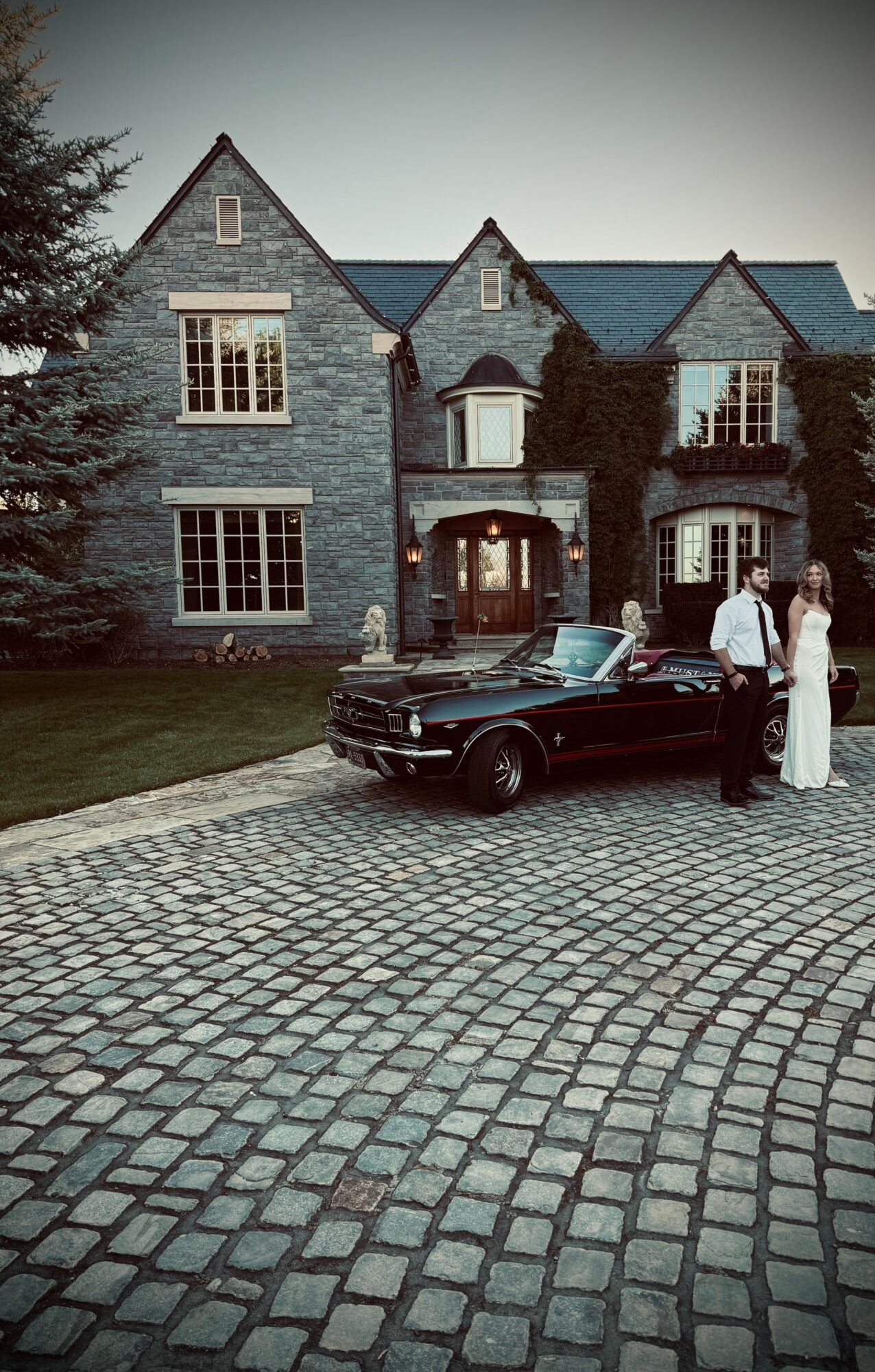 Large stone mansion with multiple gables and windows, a cobblestone driveway, a black car, and two people standing outside.