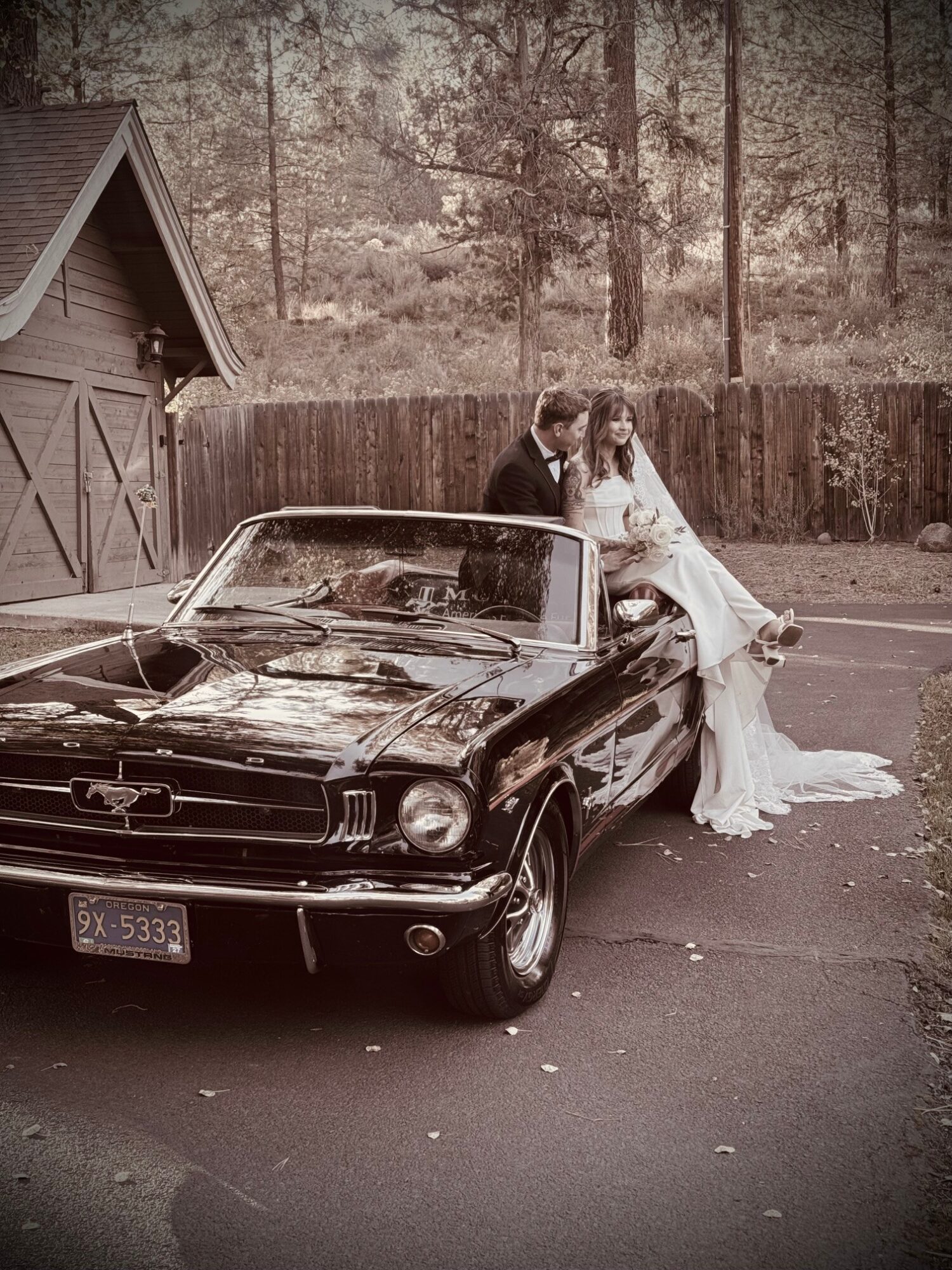 A couple in wedding attire sitting on a vintage black car parked outdoors, with a wooden fence and trees in the background.