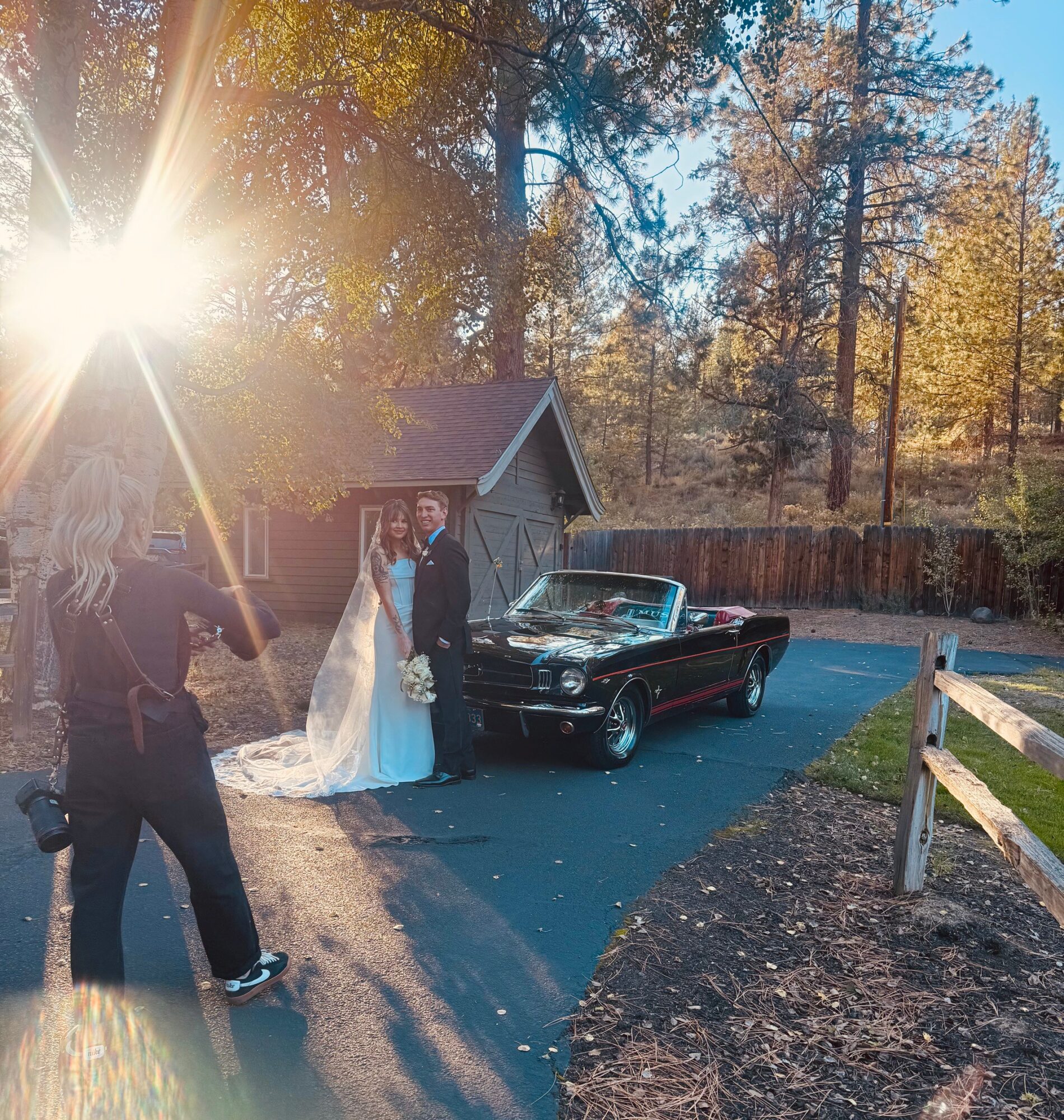 Bride and groom stand next to a black convertible car outdoors, with a photographer taking their picture, sunlight shining through trees.