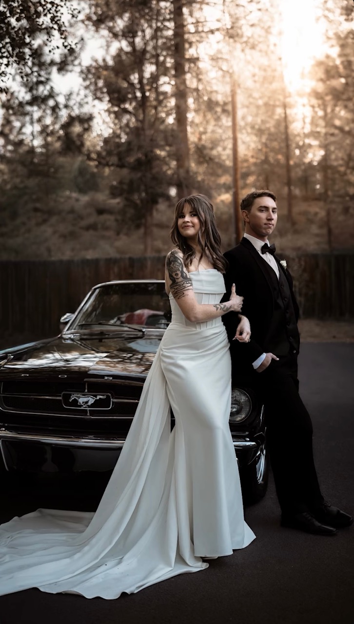 A bride and groom stand in front of a black car outdoors, with trees and sunlight in the background.