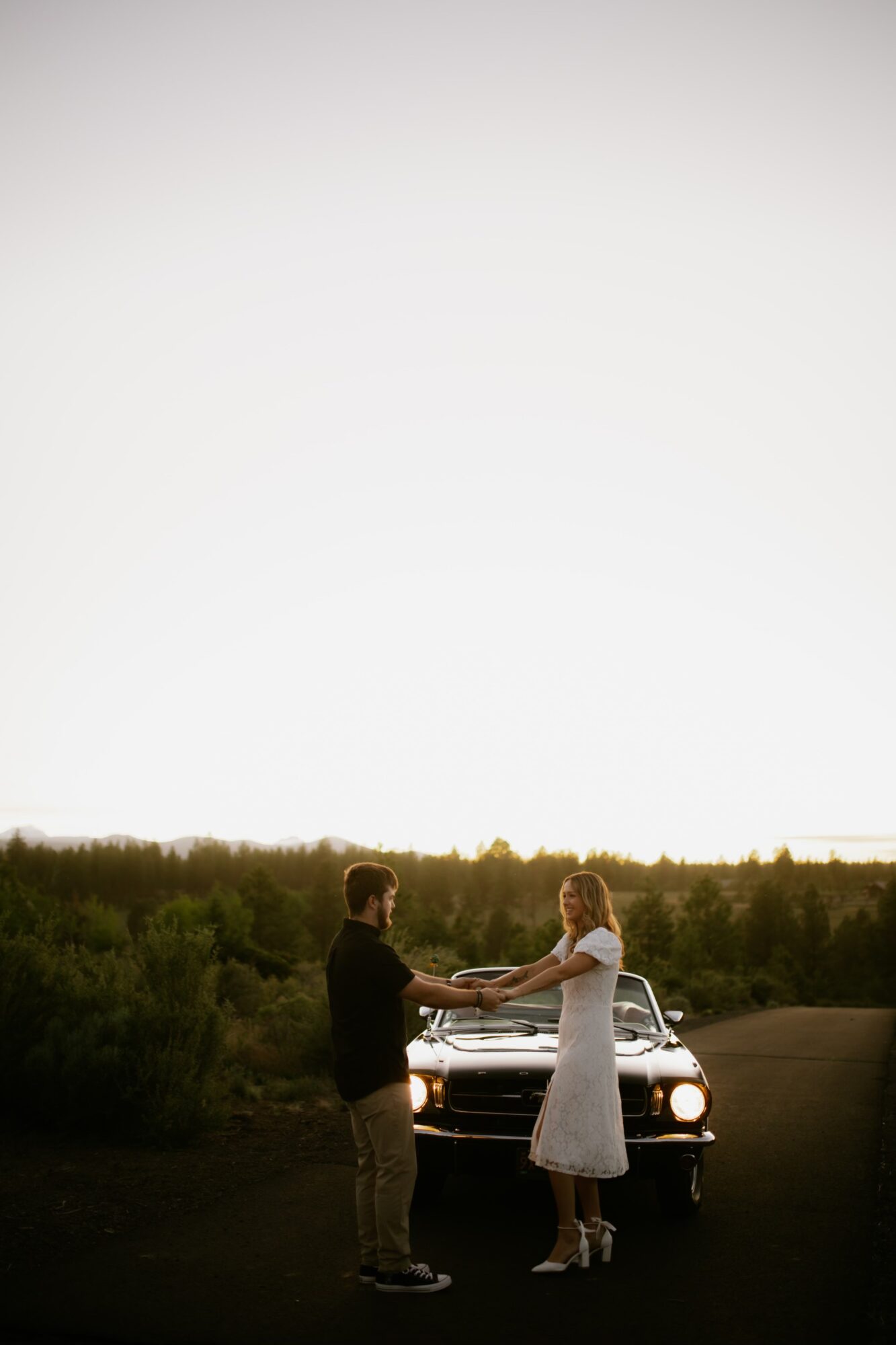 A man and woman exchange rings outdoors near a car, with a landscape and sky in the background.