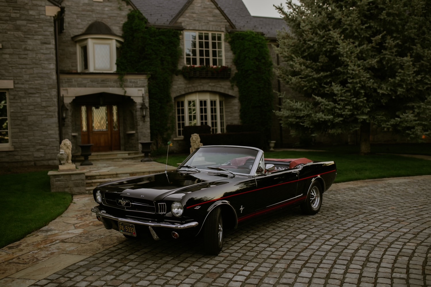 Black convertible car parked on a cobblestone driveway in front of a large house with stone walls and greenery.