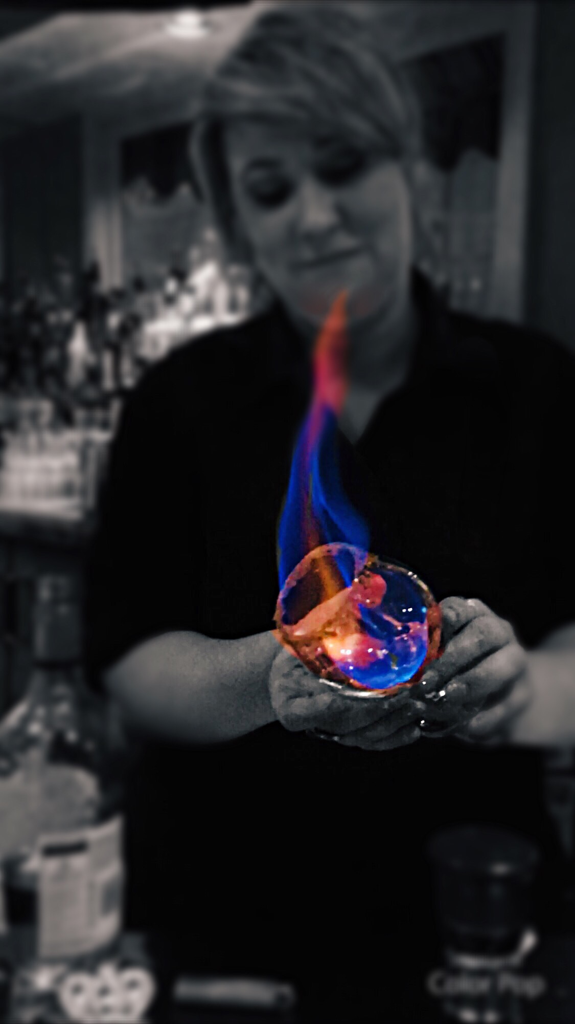 Person holding a glass with colorful flames, in a dimly lit setting, with a blurred background.