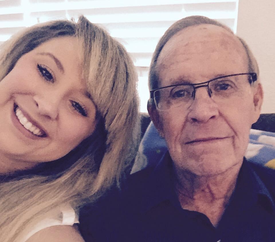 A young woman with blonde hair smiling next to an older man with glasses, both indoors with blinds behind them.