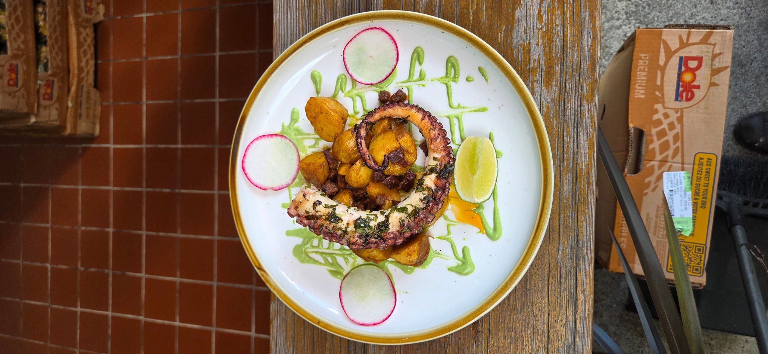 Fried octopus tentacle on a white plate with lemon, onion, and greens, on a wooden table.