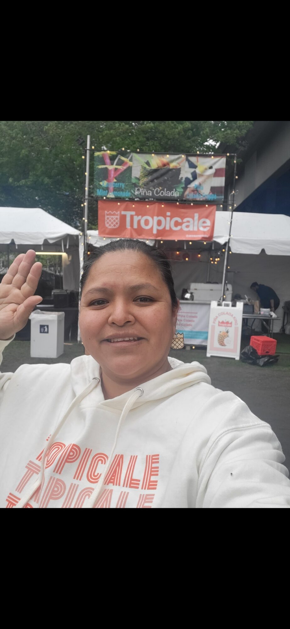 Person taking selfie outdoors, smiling, with a tent and a sign reading 'Tropicale' in the background.