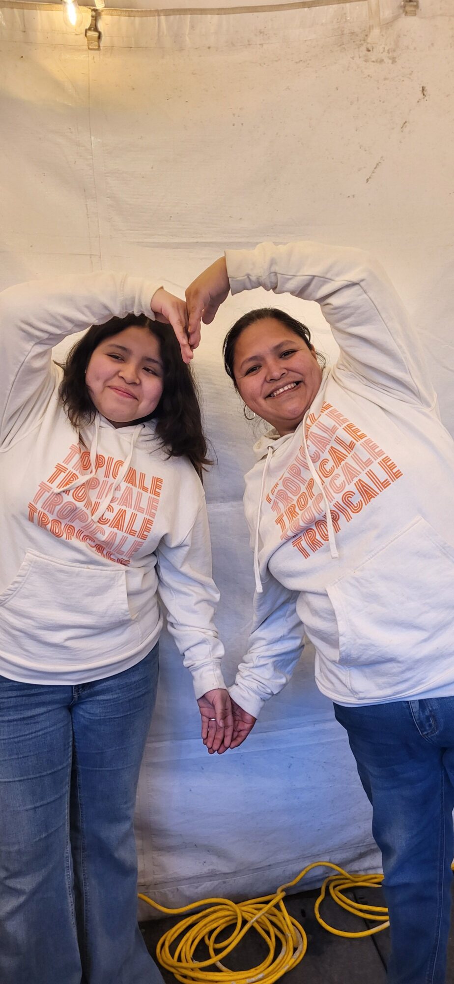 Two women lying on the floor, smiling, wearing white sweatshirts with orange text, against a plain wall background.