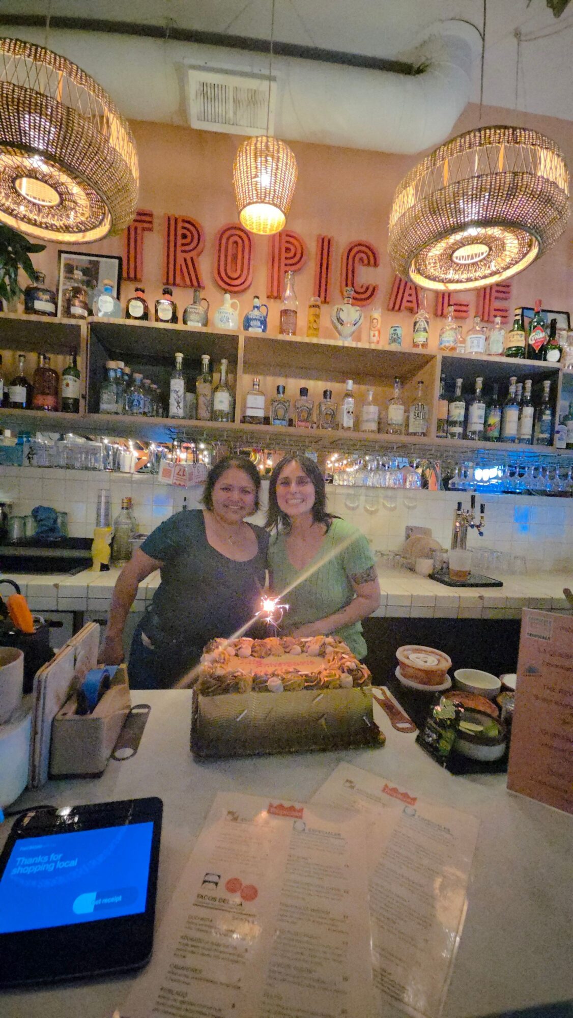 Two women stand behind a counter in a restaurant or cafe, with a menu and various items on the counter.