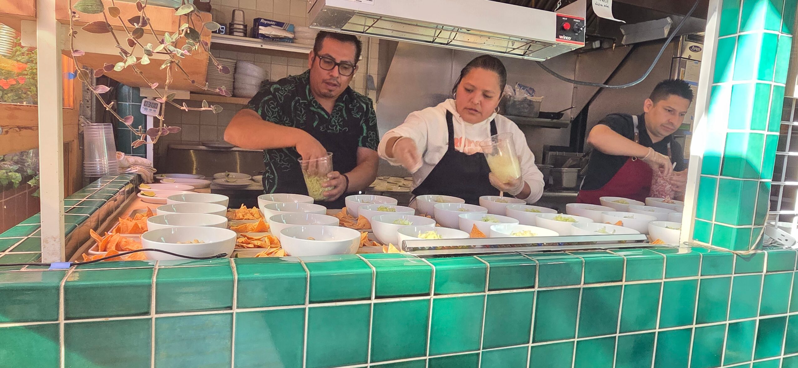 Three people working behind a food counter with green tiled front, preparing food with bowls and containers.