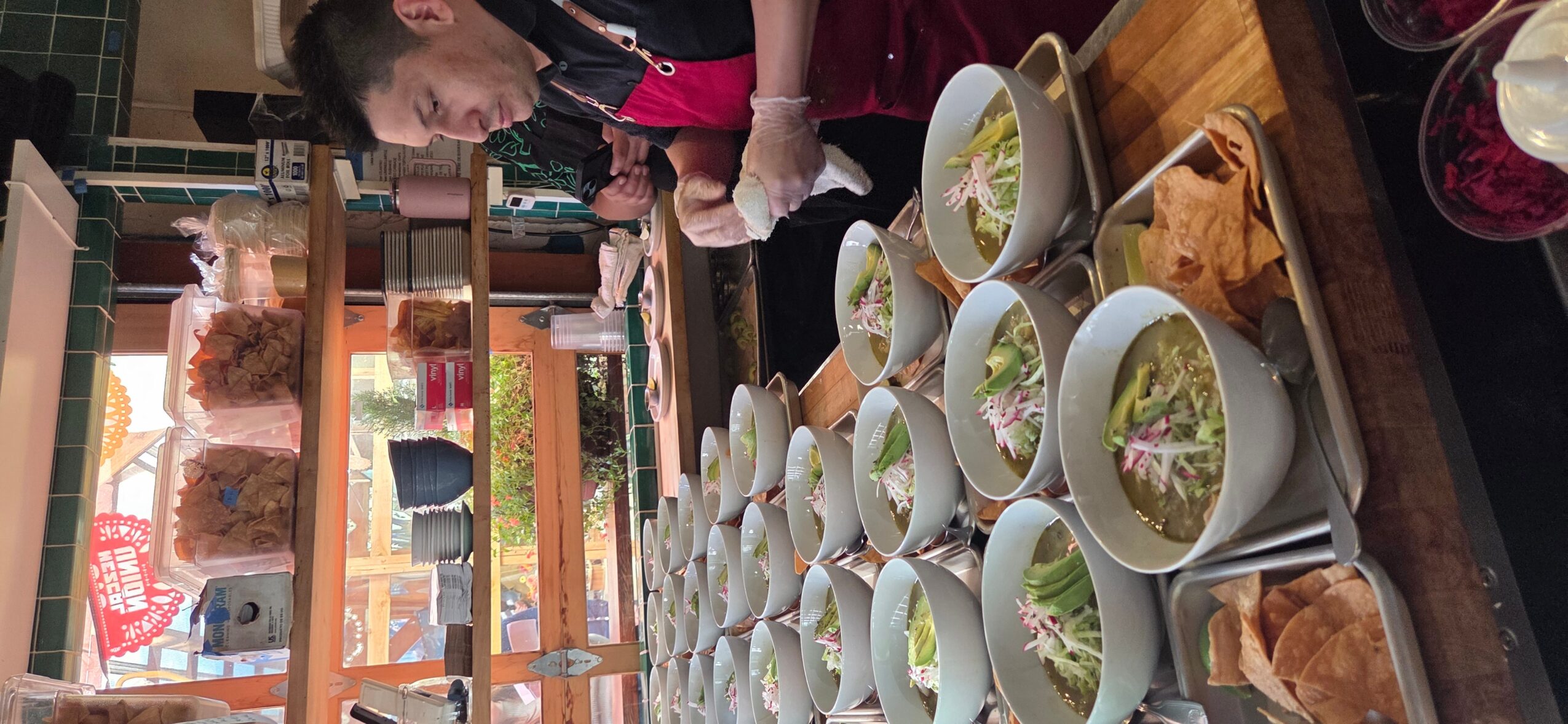 People gather around a table with bowls of salad and dishes, indoors with shelves and windows in the background.