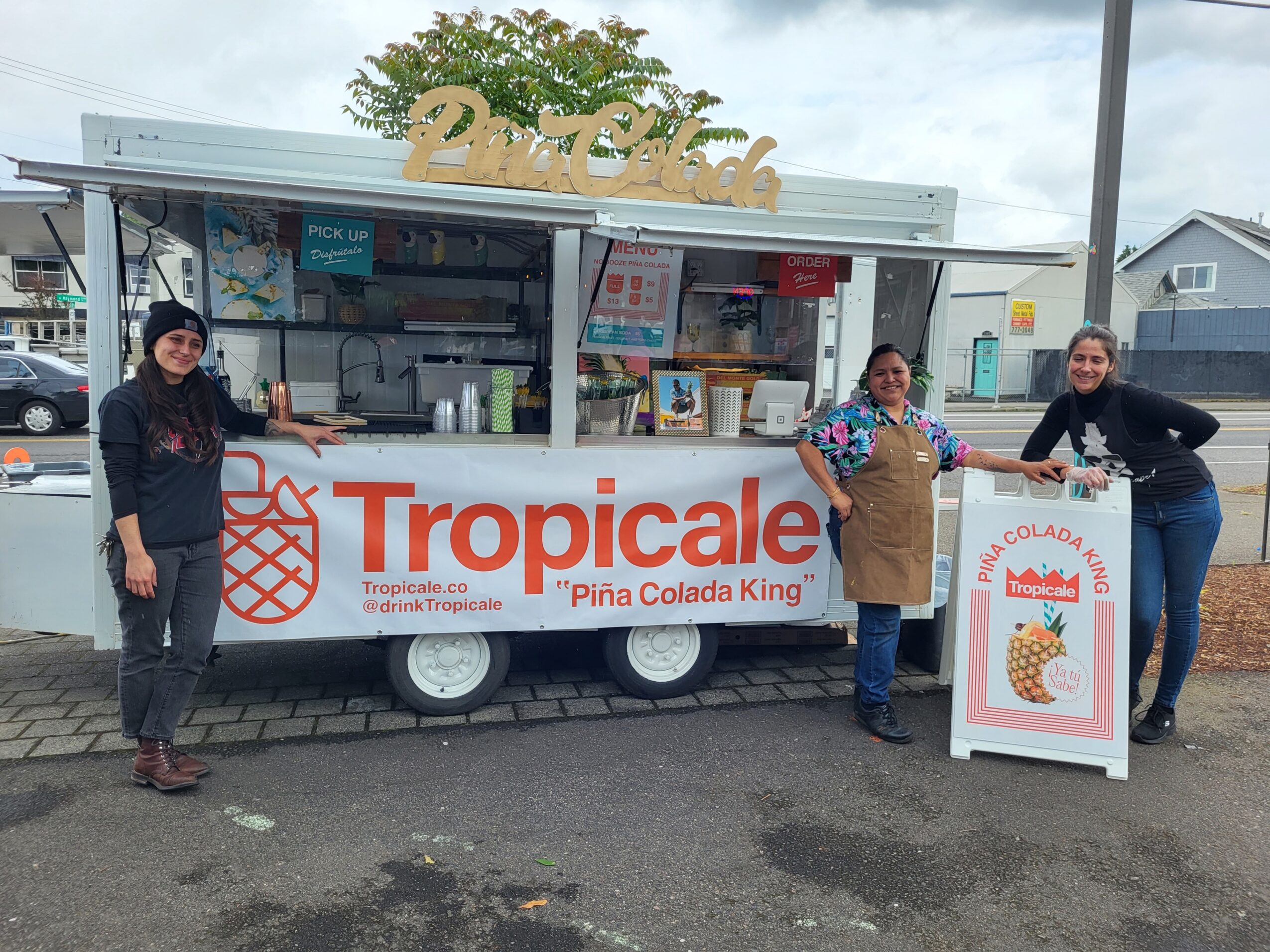 Three people stand in front of a tropical ice cream cart with a sign reading 'Tropicale Piña Colada King.'