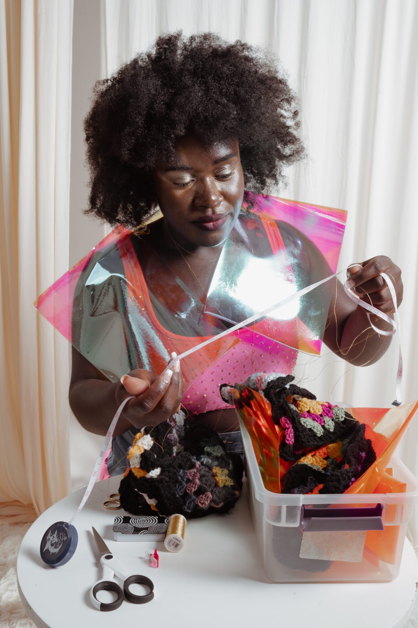 Woman with curly hair looking at colorful fabric pieces in a plastic container, sewing tools on table, curtains in background.