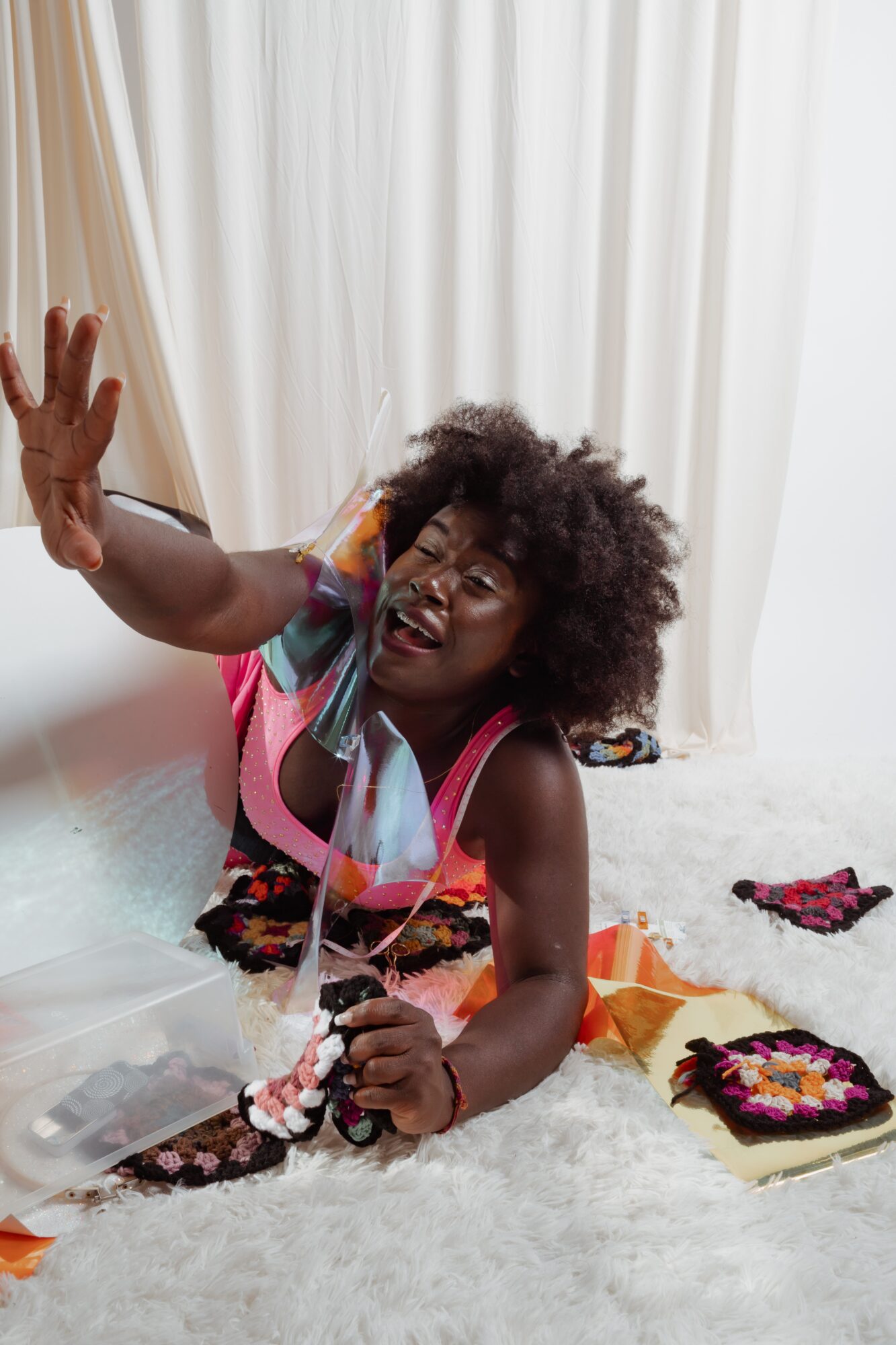 Woman with curly hair reaching out, surrounded by colorful items on a white surface, with white curtains behind.