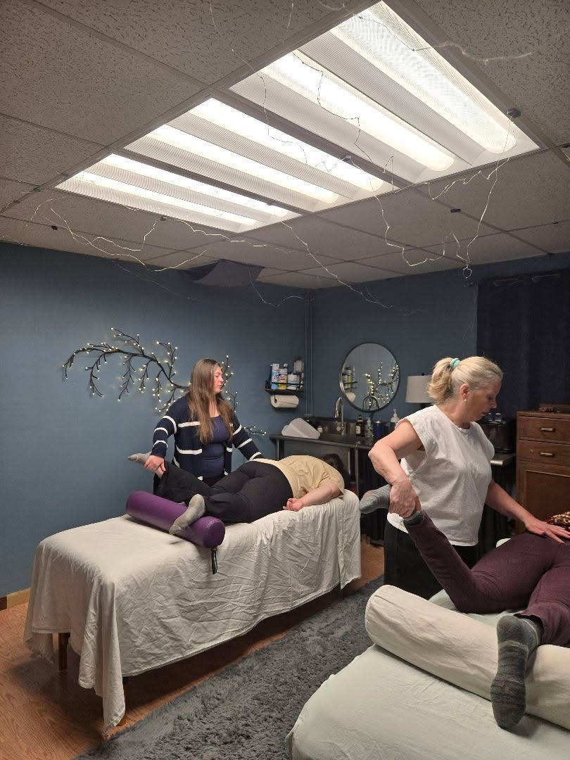 A massage room with two women, one lying face down on a massage table and the other standing nearby, in a room with blue walls and ceiling lights.