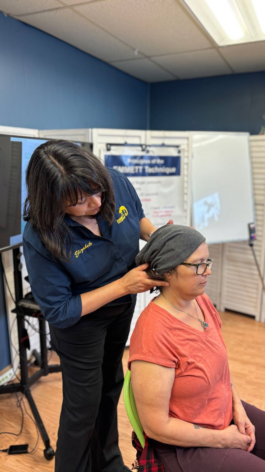Woman adjusting a head covering on a seated woman in a room with a whiteboard and blue walls.