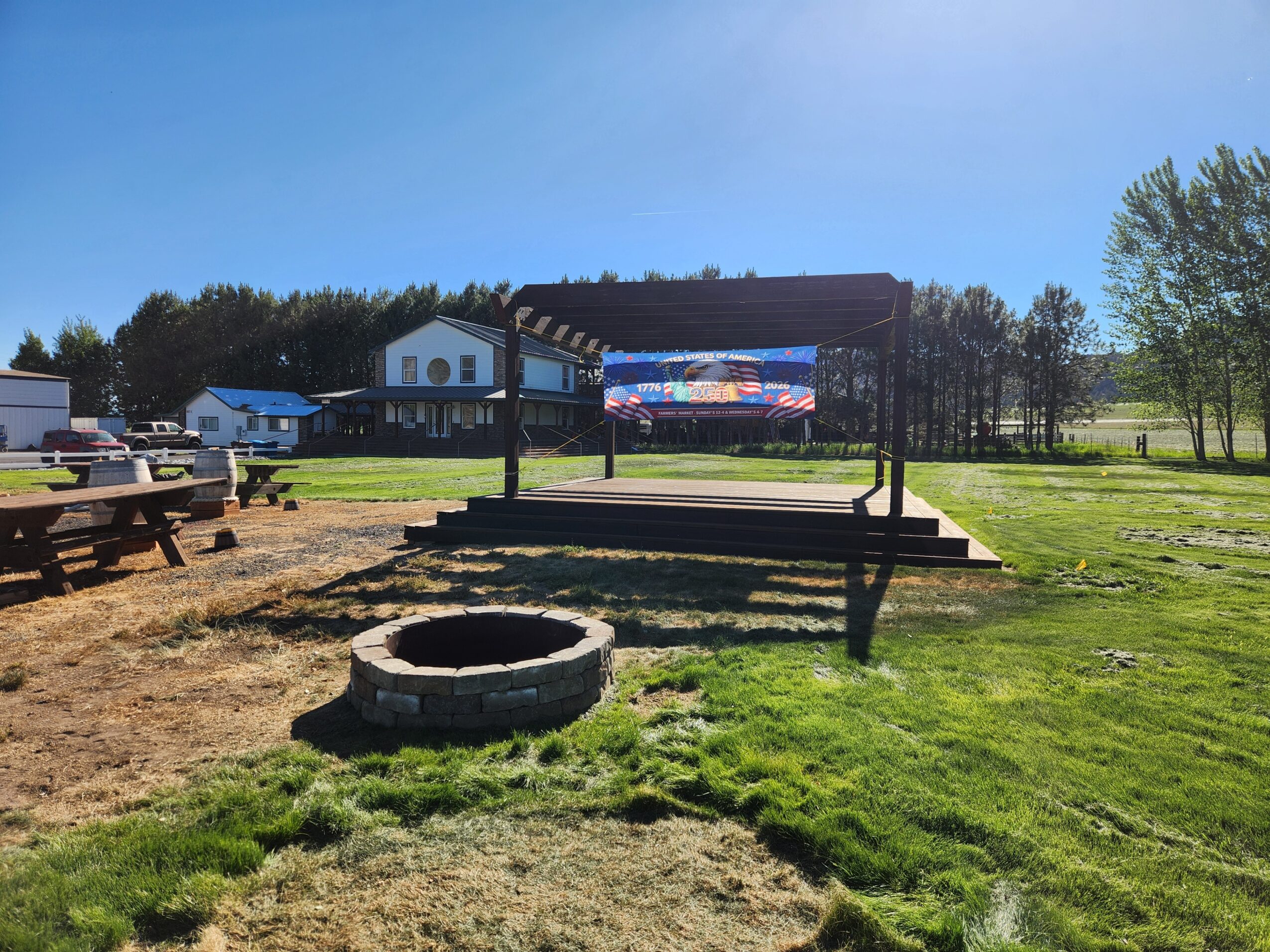 Outdoor stage with a screen, surrounded by grass, trees, and picnic tables, under a clear blue sky.