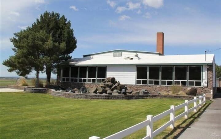 Single-story building with large windows, surrounded by a lawn, trees, and a white fence, under a blue sky.