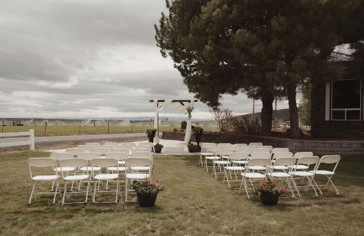 Outdoor wedding setup with chairs arranged in front of an arch, trees, and cloudy sky, with potted plants nearby.