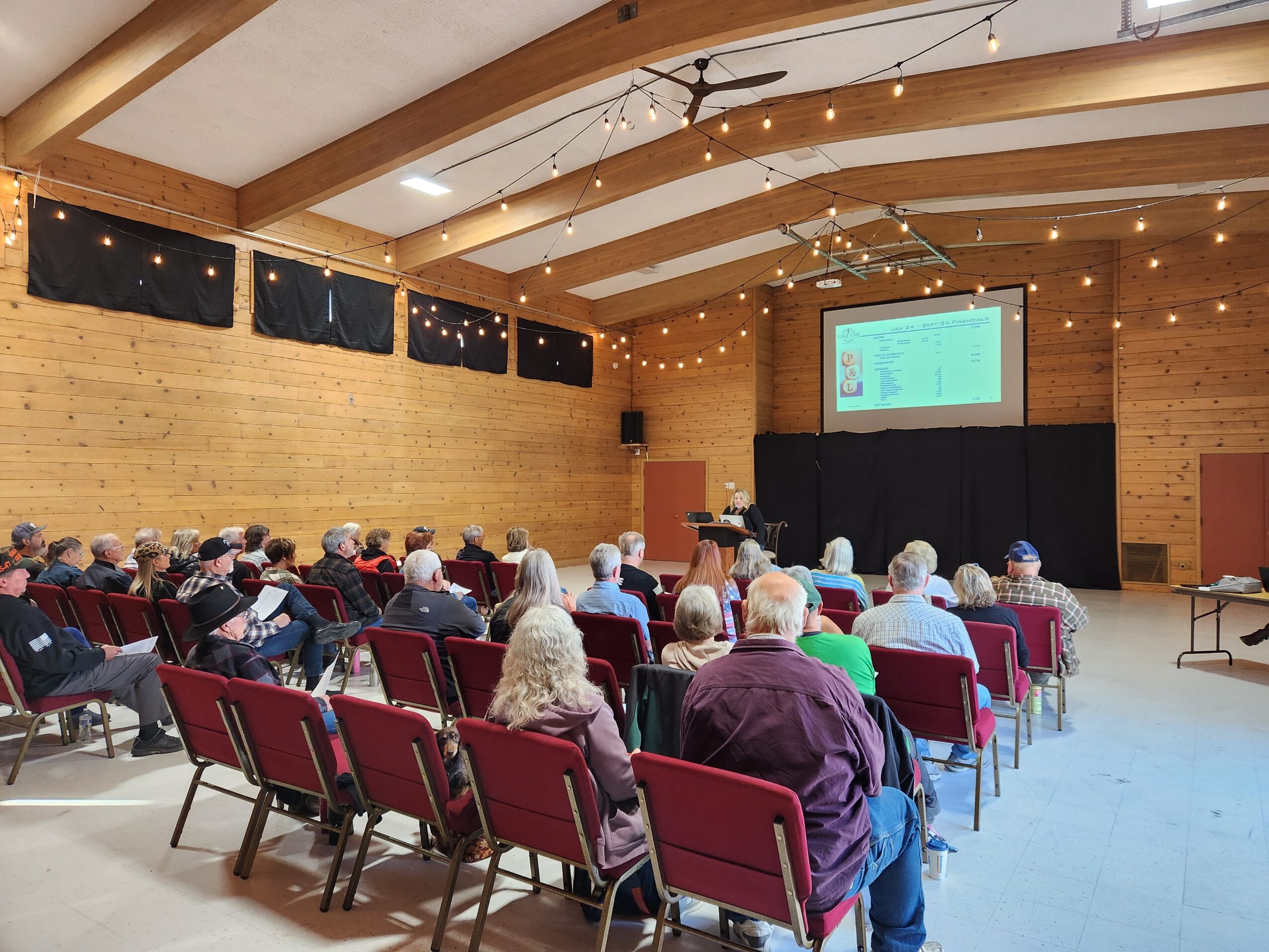Audience seated in a wooden auditorium watching a presentation on a large screen, with string lights overhead.