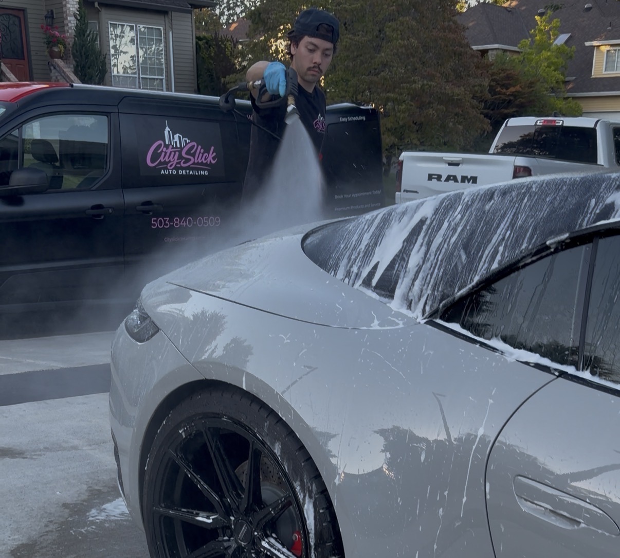 Person washing a silver car with foam, parked outdoors in a residential area, with other vehicles and houses in background.