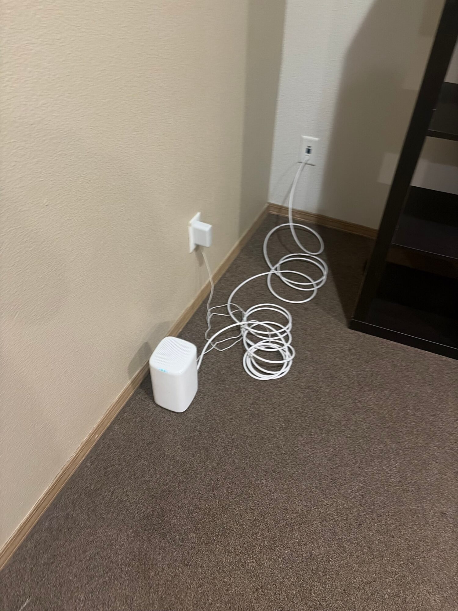 Cables and a white device on the floor near a wall and carpet, with a power outlet and a black shelf above.