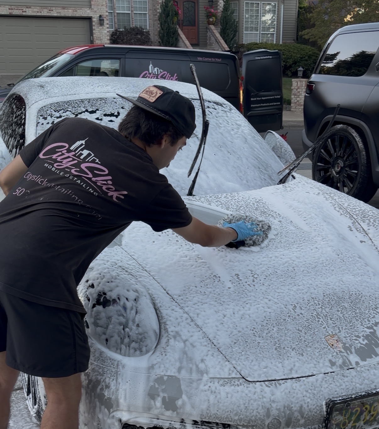 Person washing a car with soap and sponge outdoors, other vehicles and houses in background.