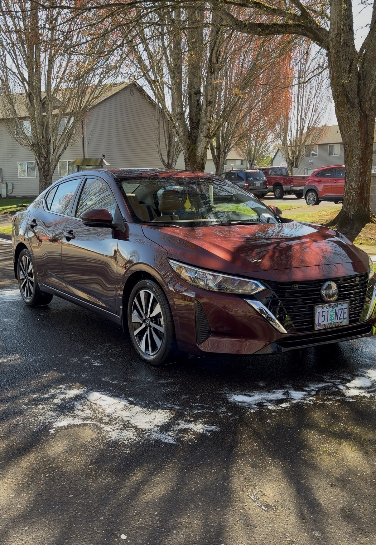 Red sedan parked on a street with trees and houses in the background, sunlight casting shadows on the ground.