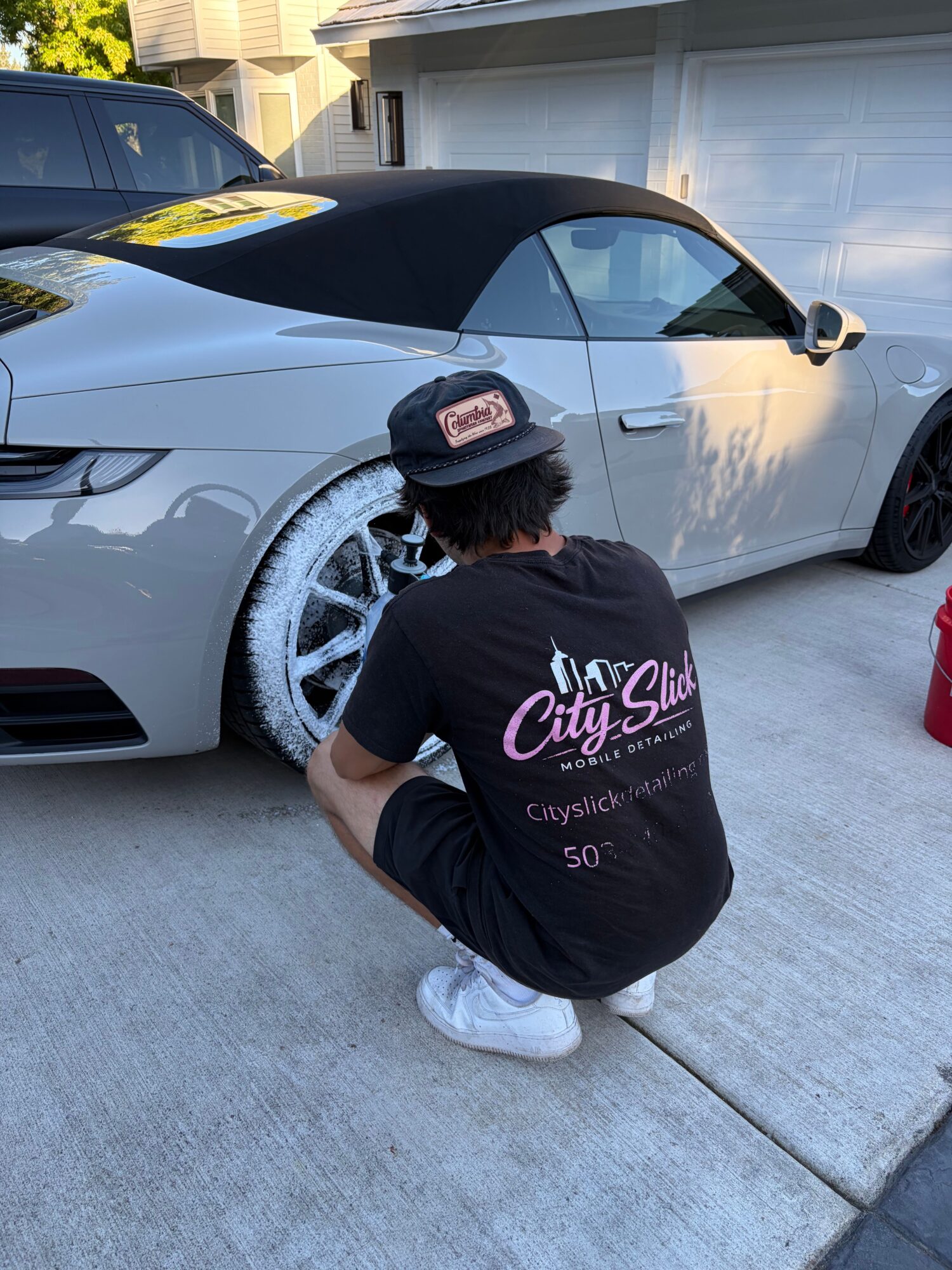 Person crouching next to a car's front wheel, cleaning or inspecting it, on a driveway.