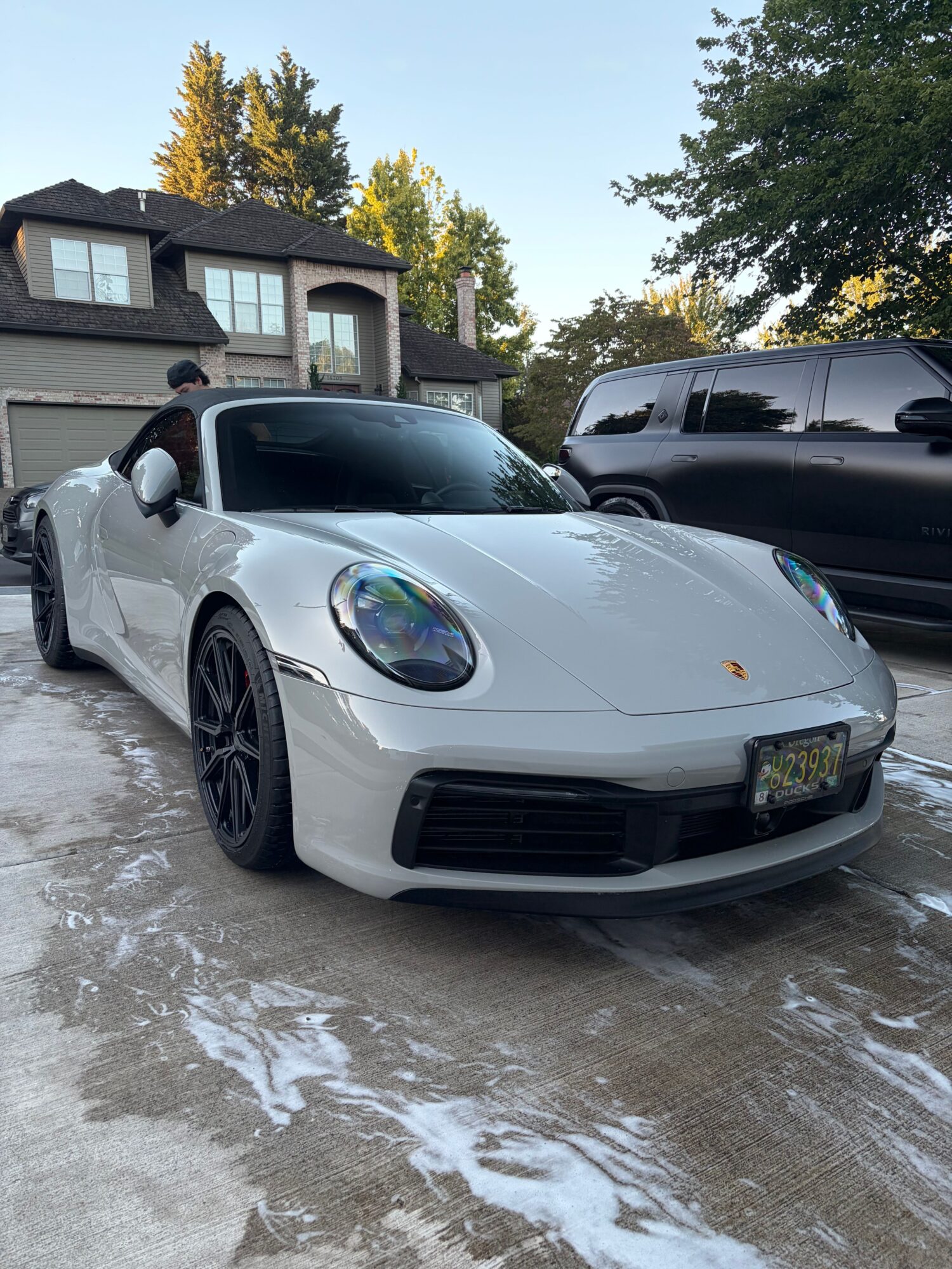 White sports car parked on wet ground with trees and buildings in background.