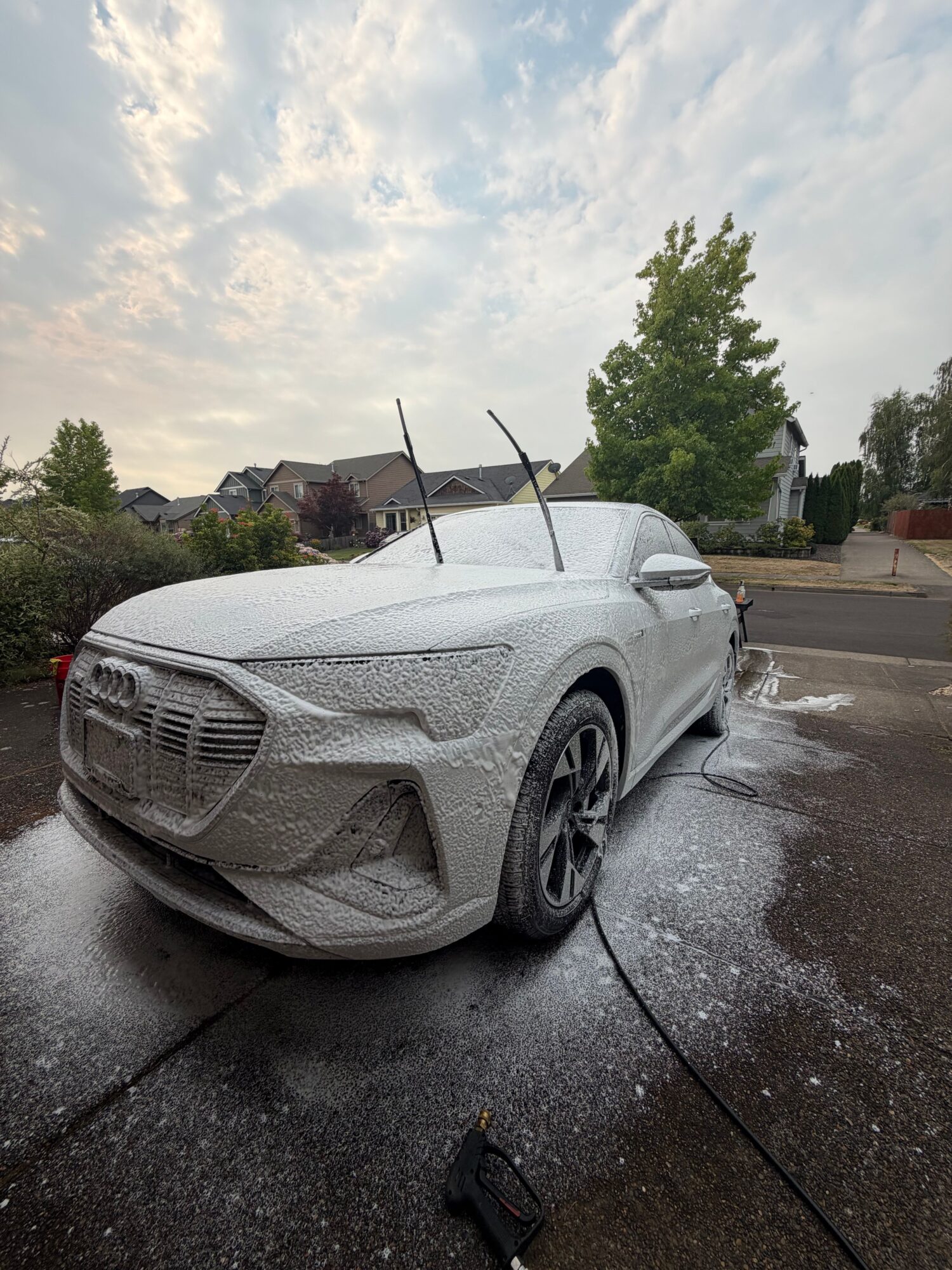 White sports car parked on wet driveway with trees and houses in background.