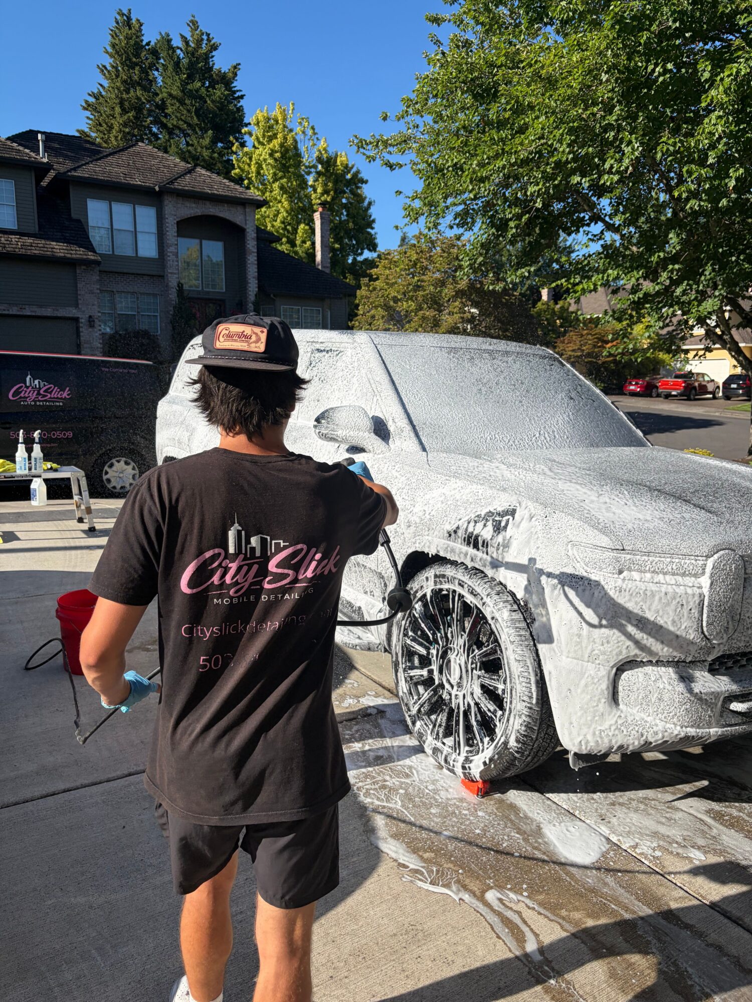 Person in black dress with pink text, washing a silver car outdoors on a sunny day.