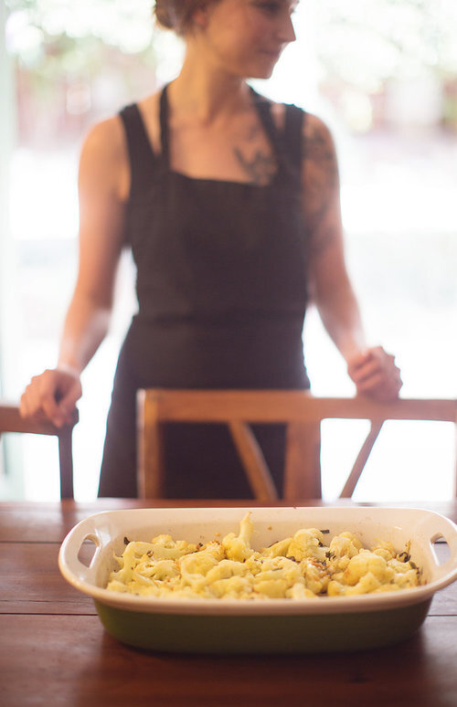 Person standing behind a table with a dish of pasta, blurred background, natural light, wooden chairs, indoor setting.