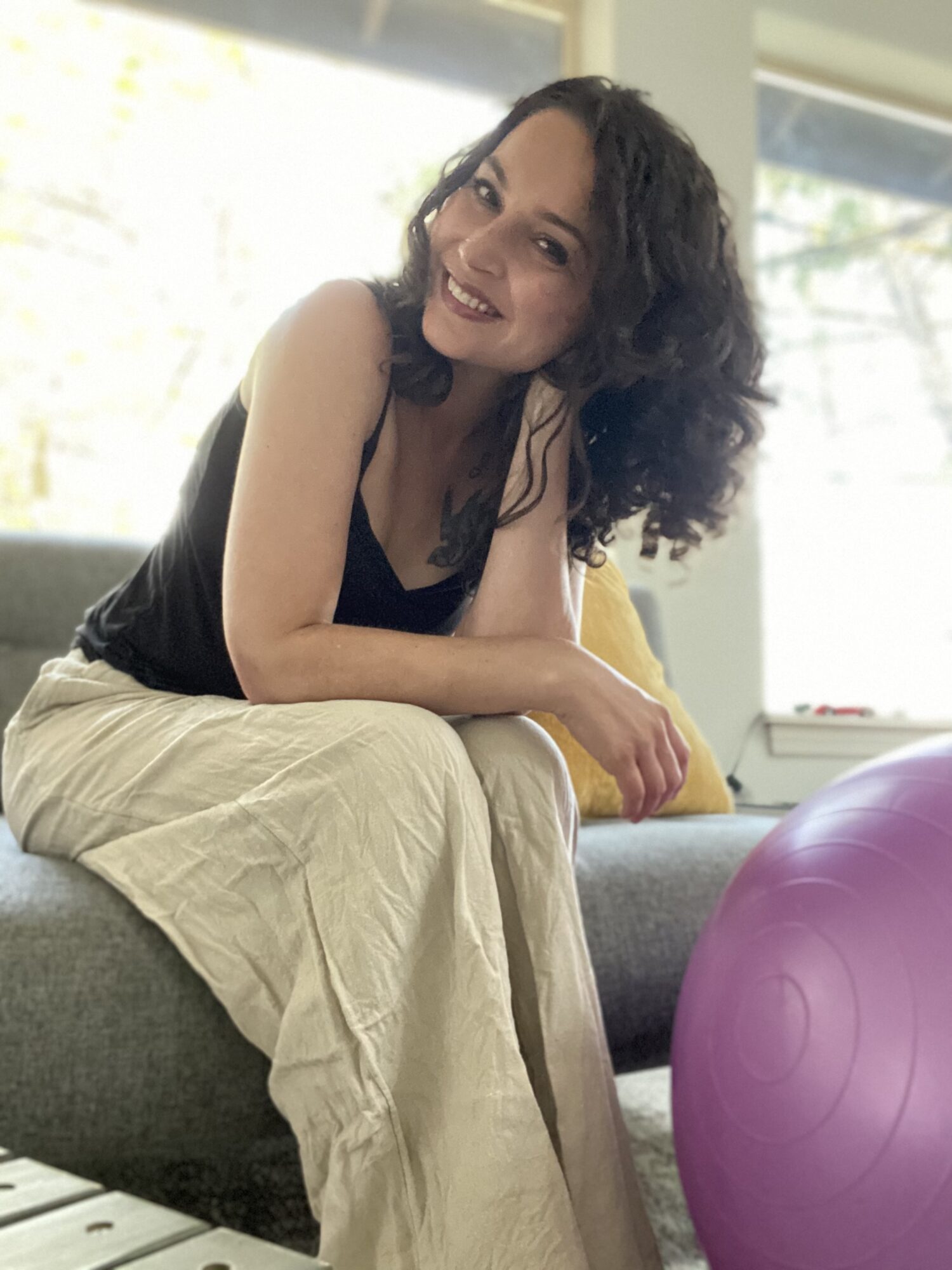 Woman with curly hair smiling, sitting on a couch indoors, with a purple exercise ball nearby.