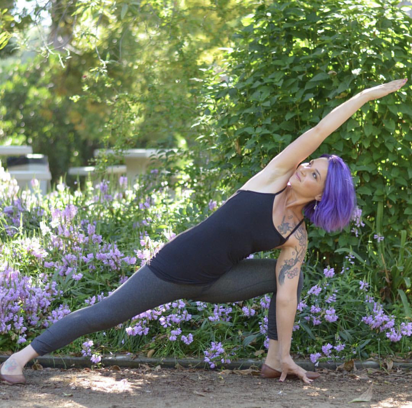 Woman with purple hair in a yoga pose outdoors, stretching with one arm extended over her head, surrounded by flowers and greenery.