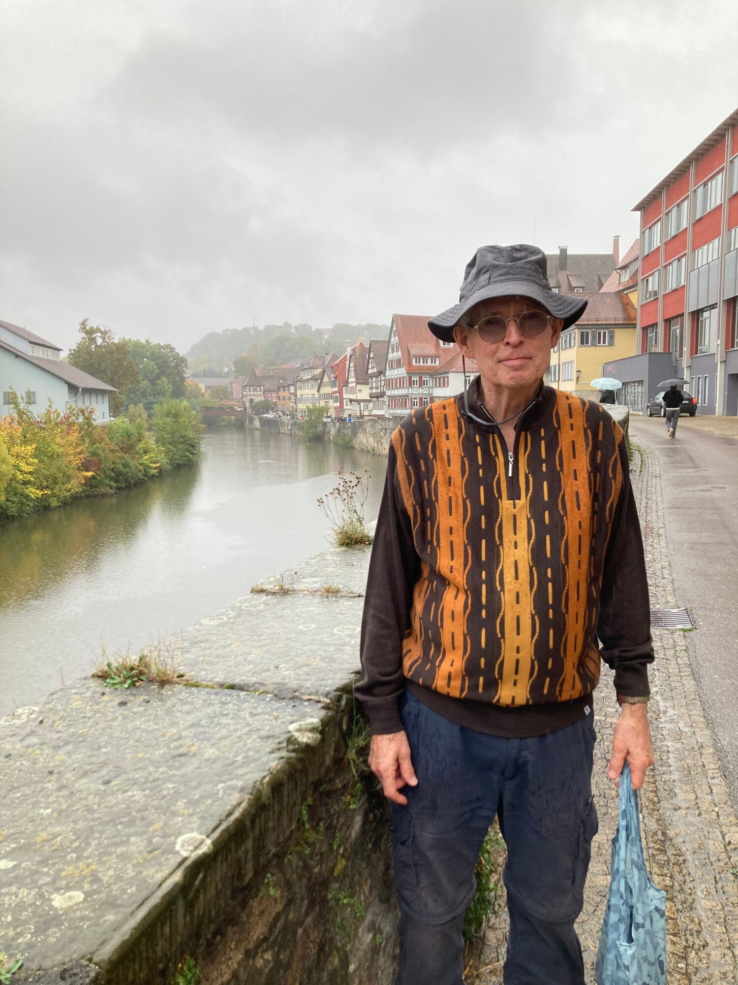 Person standing on a canal bank with buildings and cloudy sky in the background.