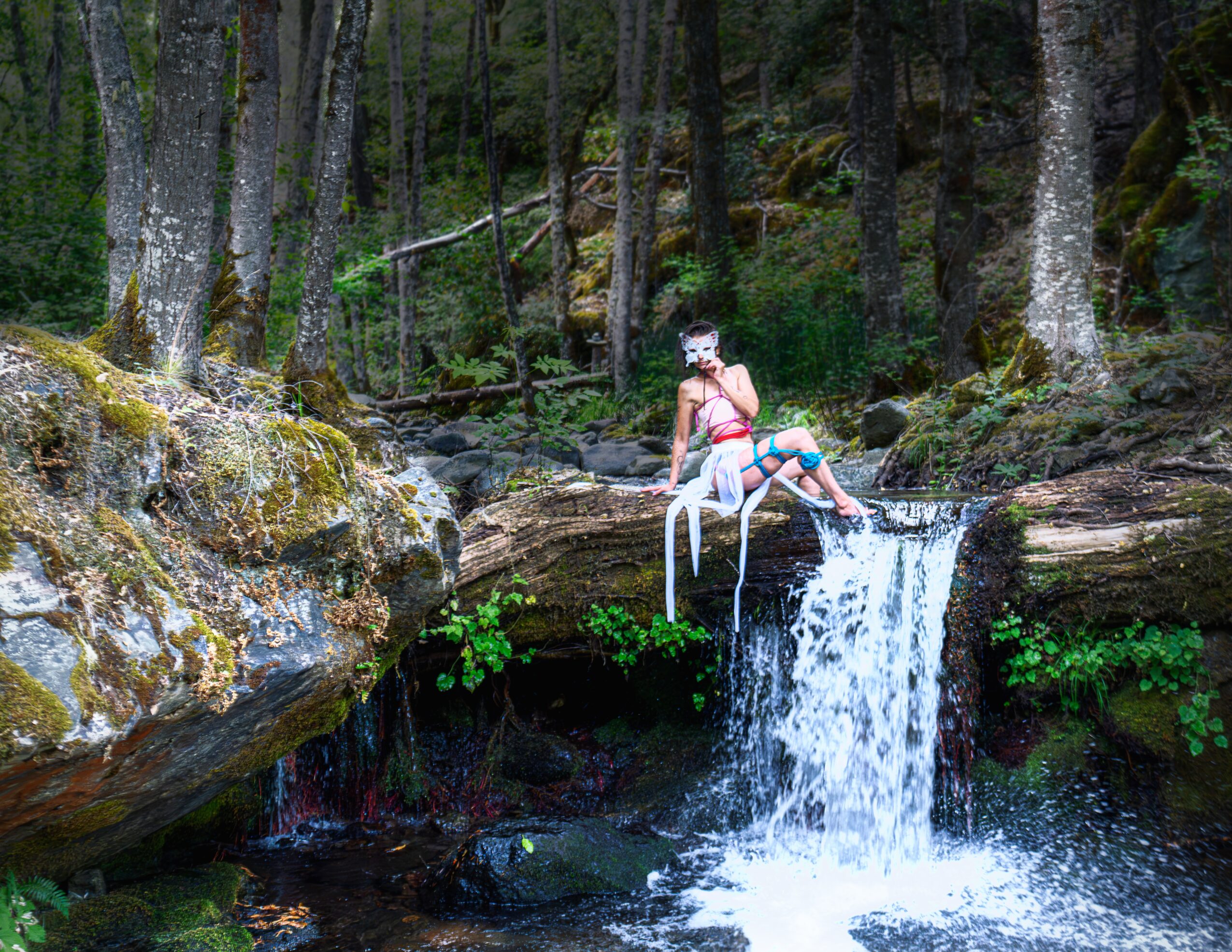 Child sitting on a rock above a waterfall in a forest, with trees and moss around.