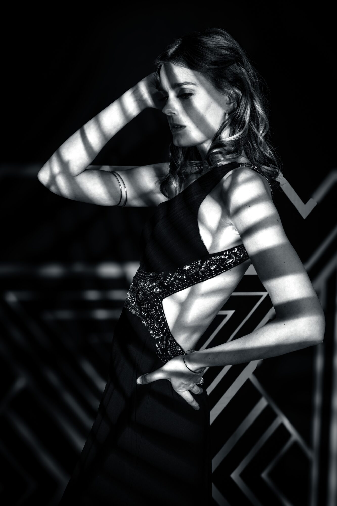 Woman with wavy hair, hand on head, in a dress with light and shadow patterns, standing against a dark background.