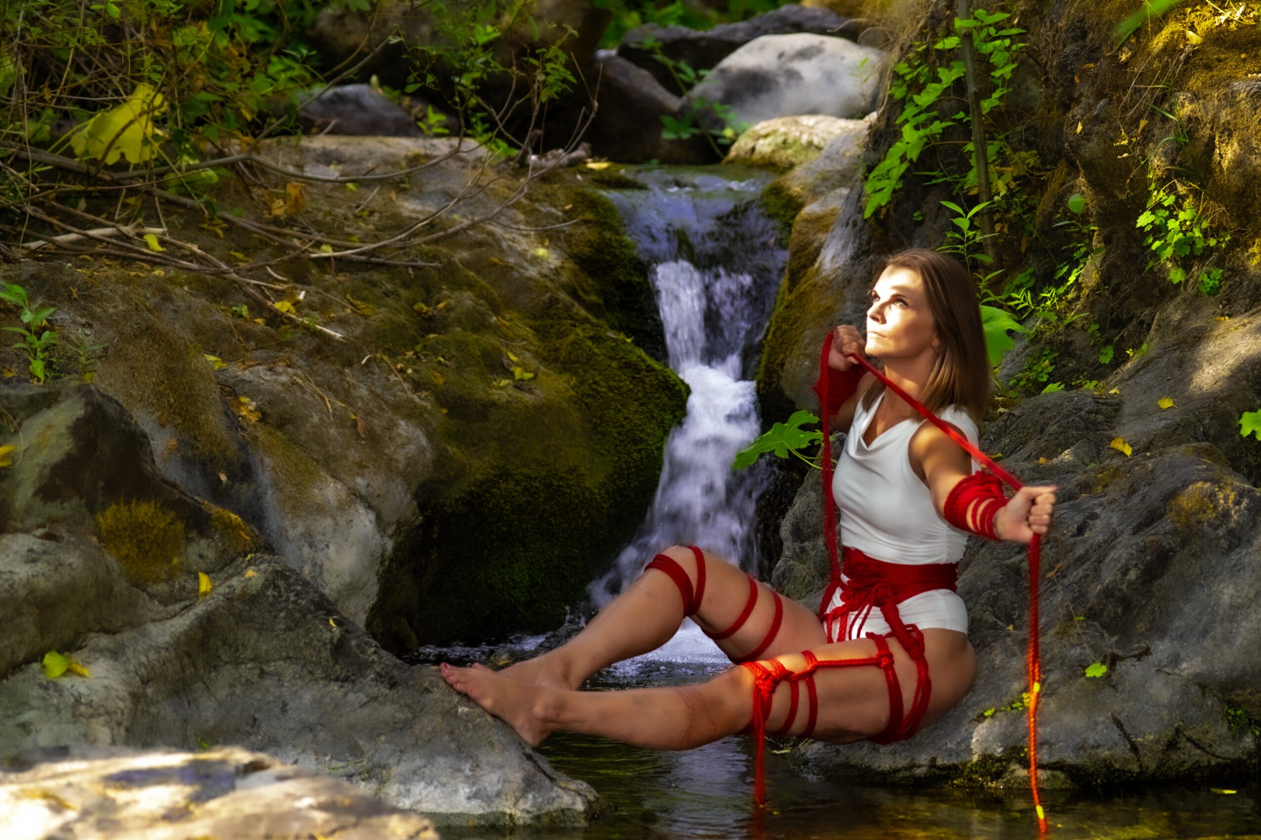 Woman sitting on rocks near a small waterfall in a forest, wearing white and red clothing, holding a red rope.