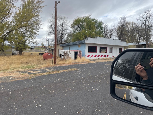 A small building with a white and red exterior, trees, and a cloudy sky in the background, seen from a roadside view.