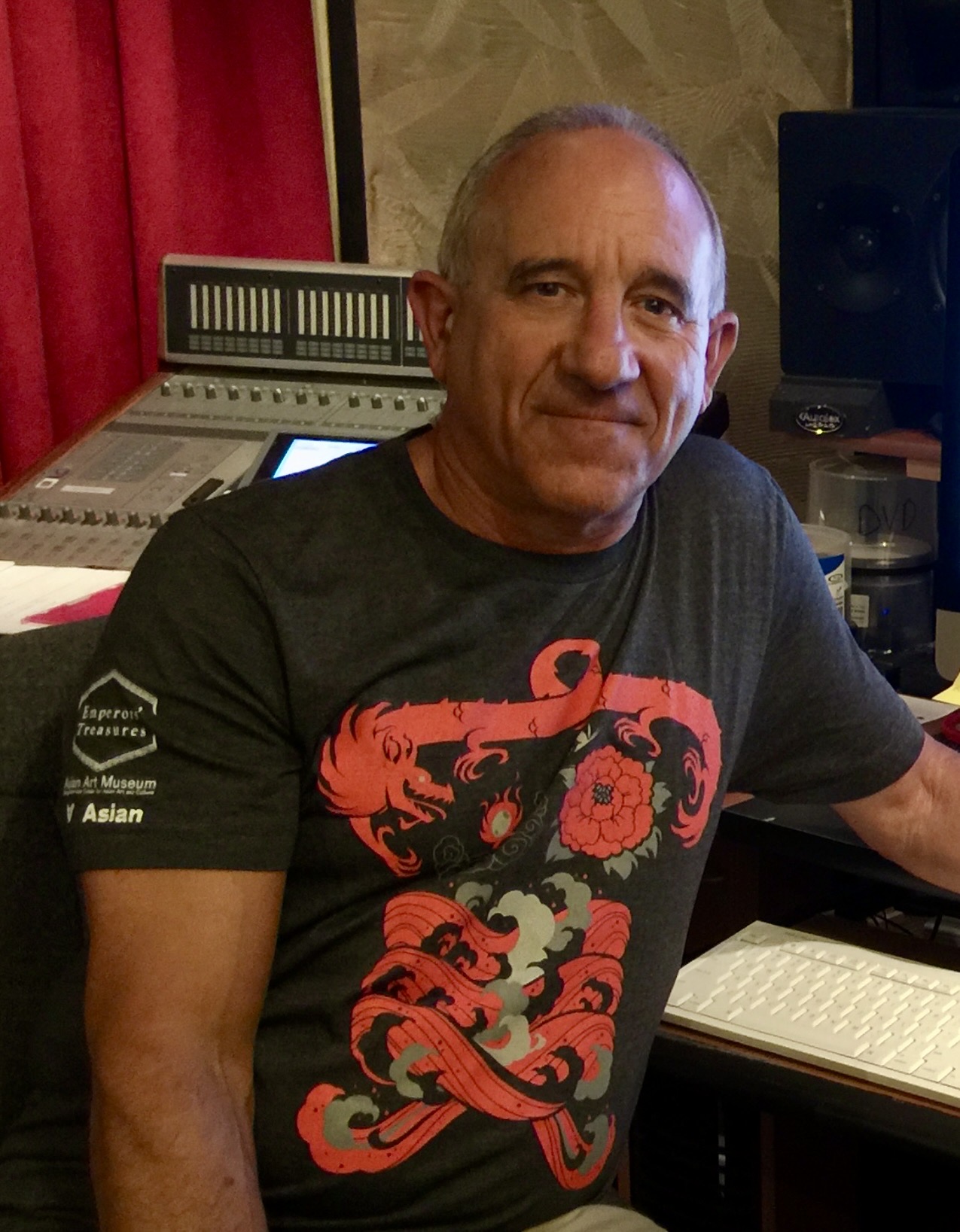 Middle-aged man with short gray hair sitting in a recording studio, wearing a black T-shirt with red and gray design, smiling.