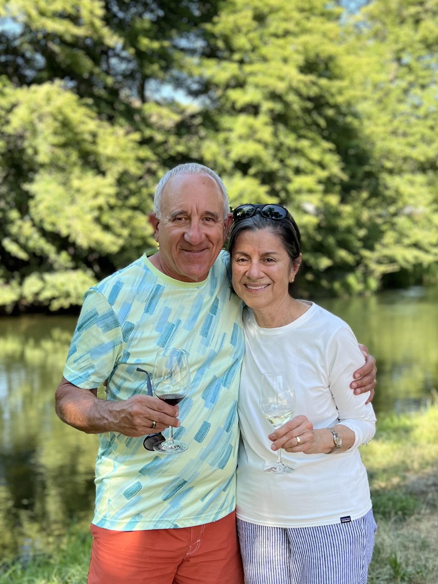 Two people smiling outdoors, holding glasses, standing close together with trees and water in background.