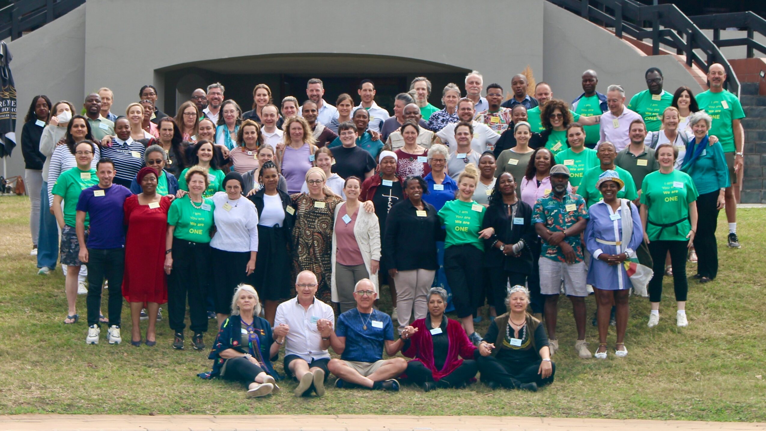 Large group of people gathered outdoors in front of a building, posing for a photo on grass.
