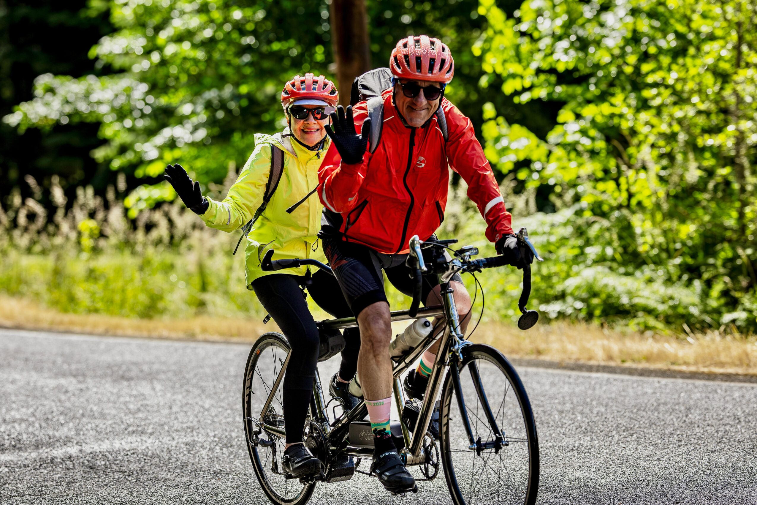Two people riding a bicycle on a road surrounded by green trees, wearing helmets and sunglasses, smiling and waving.