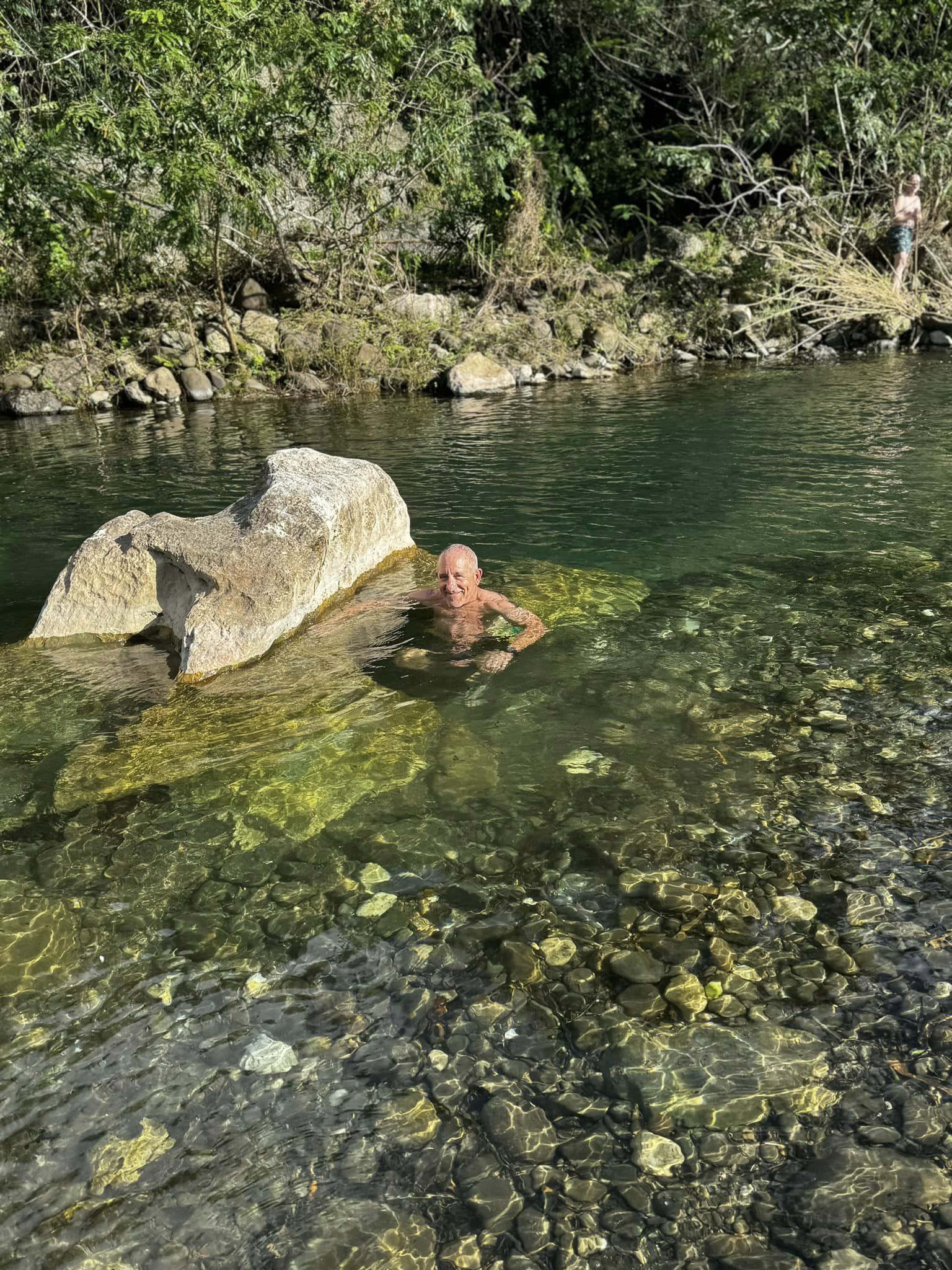 Person swimming in a clear river with rocks and green trees in the background.