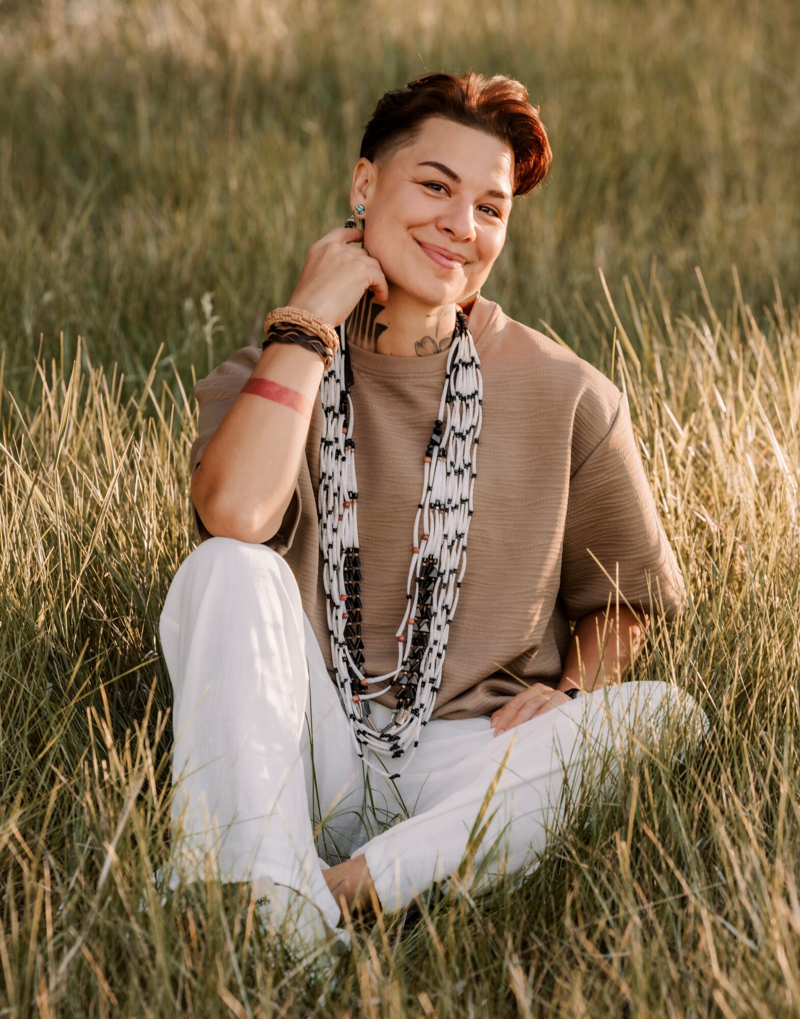 Woman sitting in grass, smiling, wearing a beige top, white pants, and a patterned scarf, outdoors in sunlight.