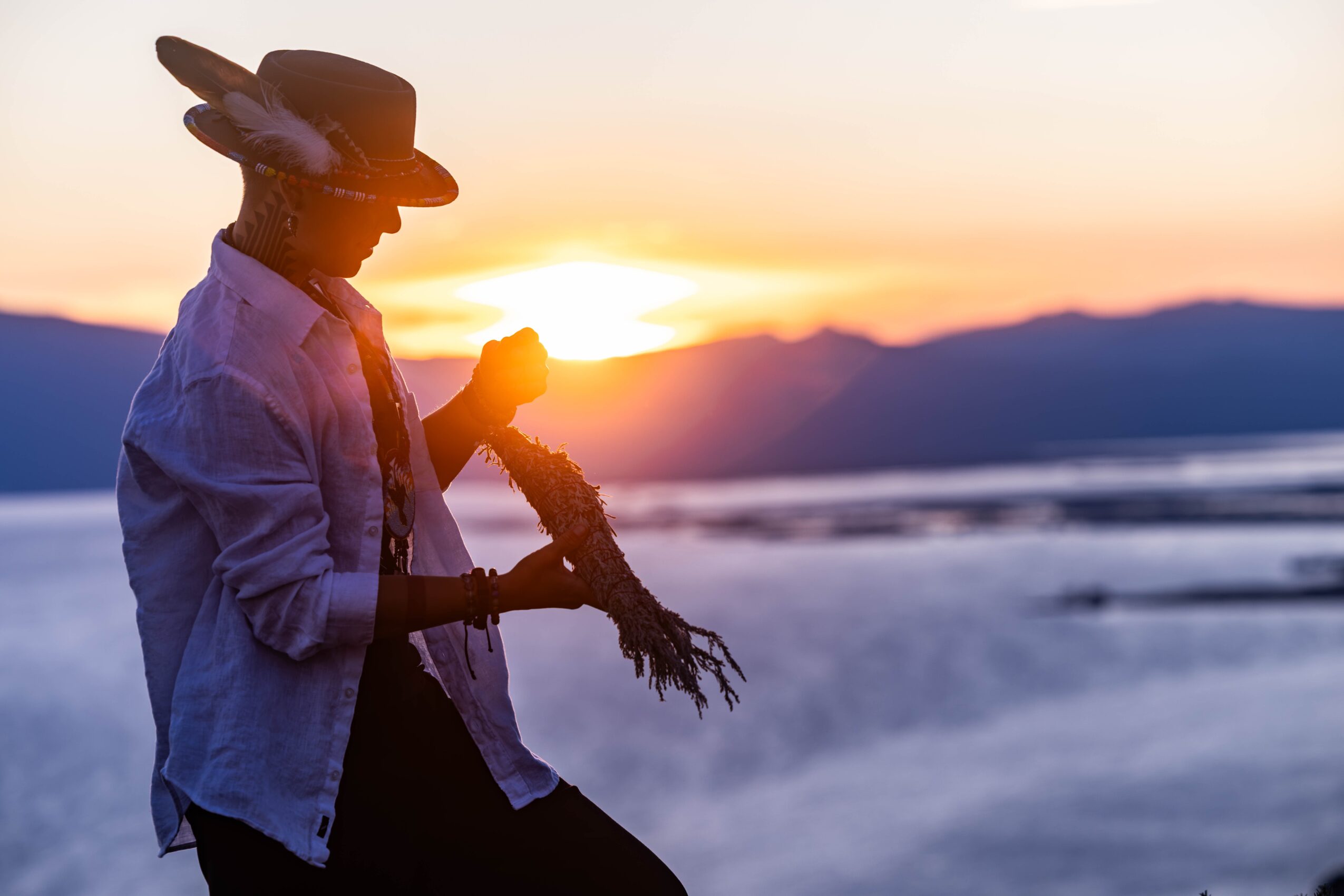 Person holding a saxophone outdoors at sunset, mountains in background, wearing a hat and casual clothing.