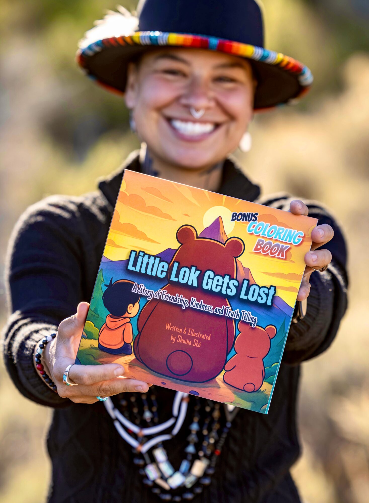 Person smiling and holding a colorful book titled 'Little Lok Gets Lost!' outdoors, wearing a hat and jewelry.