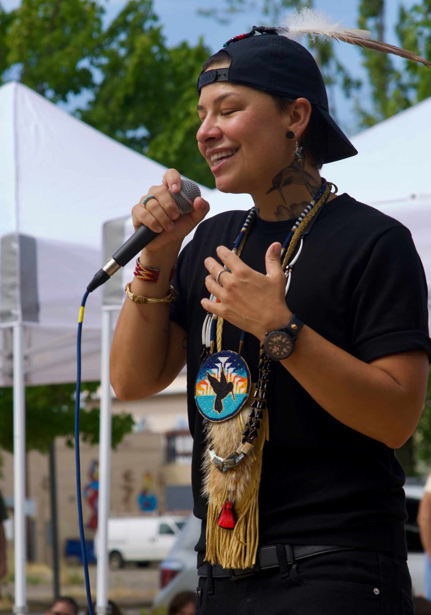 Person wearing a black cap and shirt, holding a microphone, smiling, outdoors with tents and trees in background.